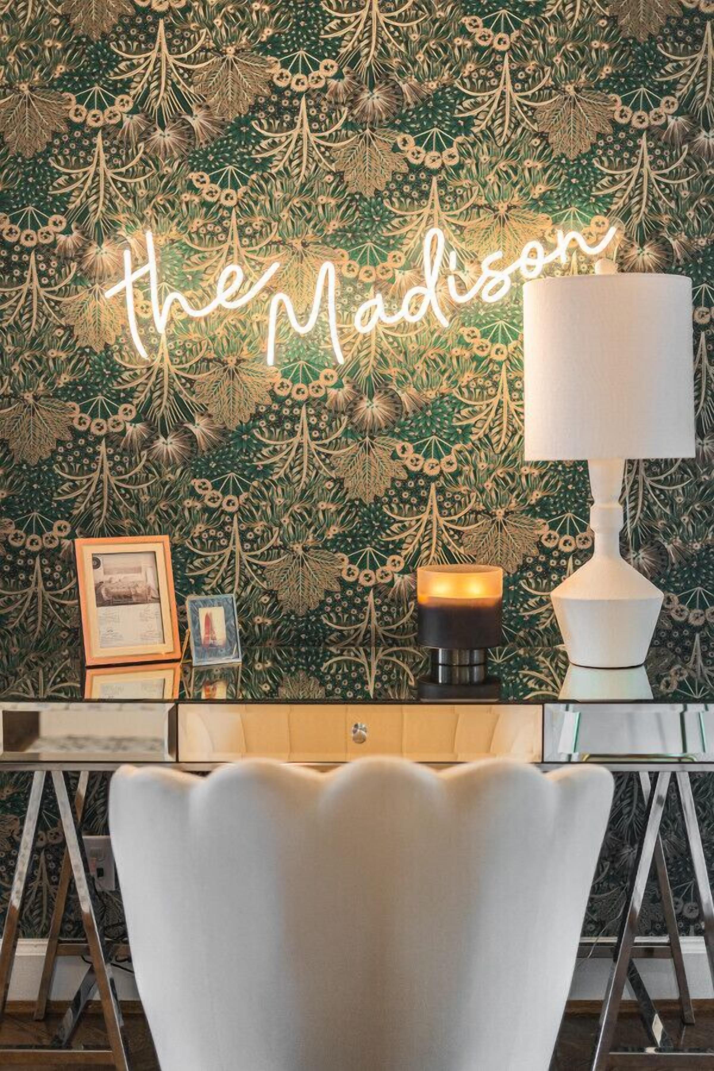 Una habitación con papel tapiz verde estampado, un letrero de neón que dice "the Madison", una lámpara de mesa blanca y una silla blanca.