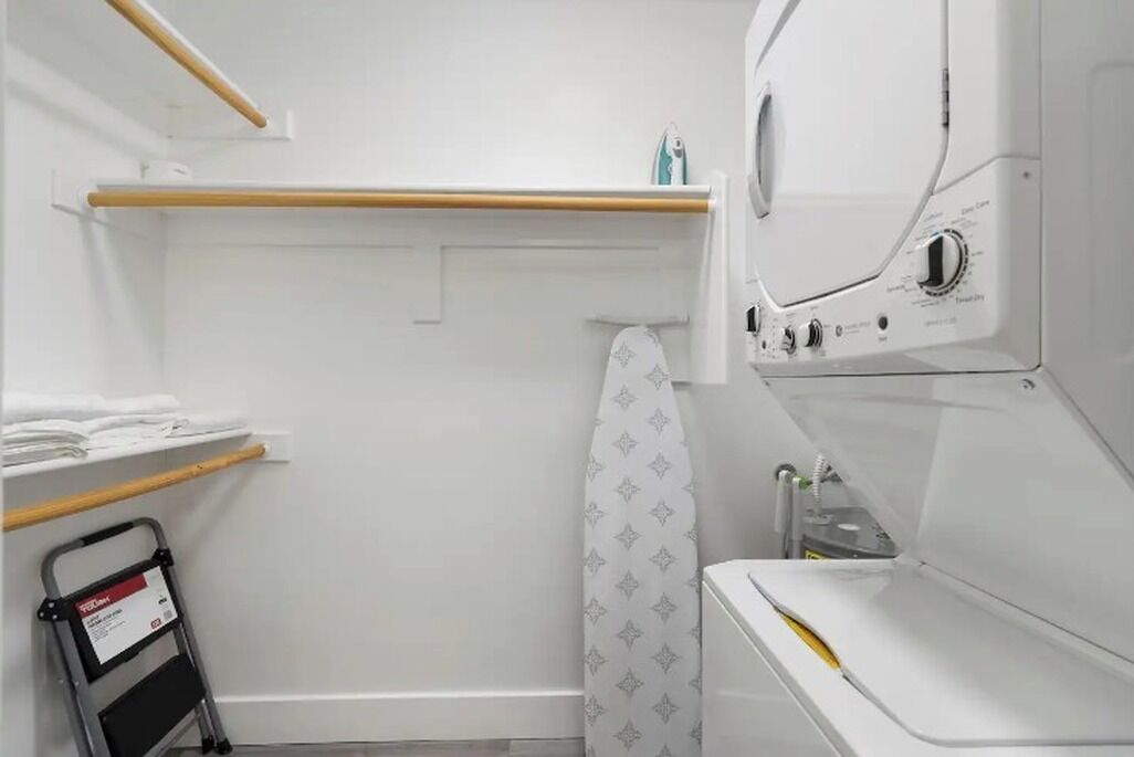 A laundry room with a washing machine, dryer, and ironing board. There is a shelf with folded clothes and a step ladder.