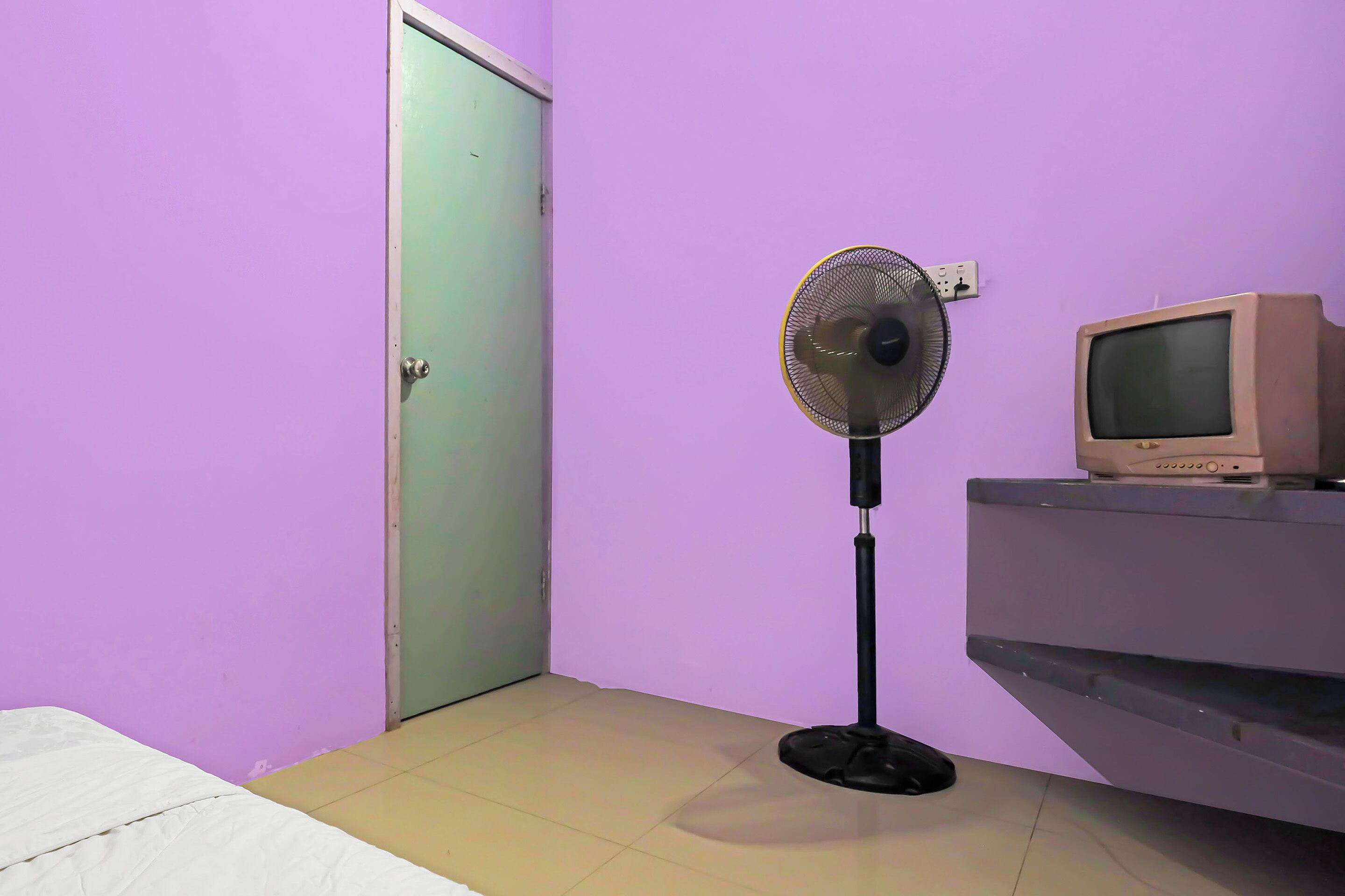 Ein Zimmer mit einem Bett, einem Standventilator, einem Fernseher auf einem Regal und einer Tür.