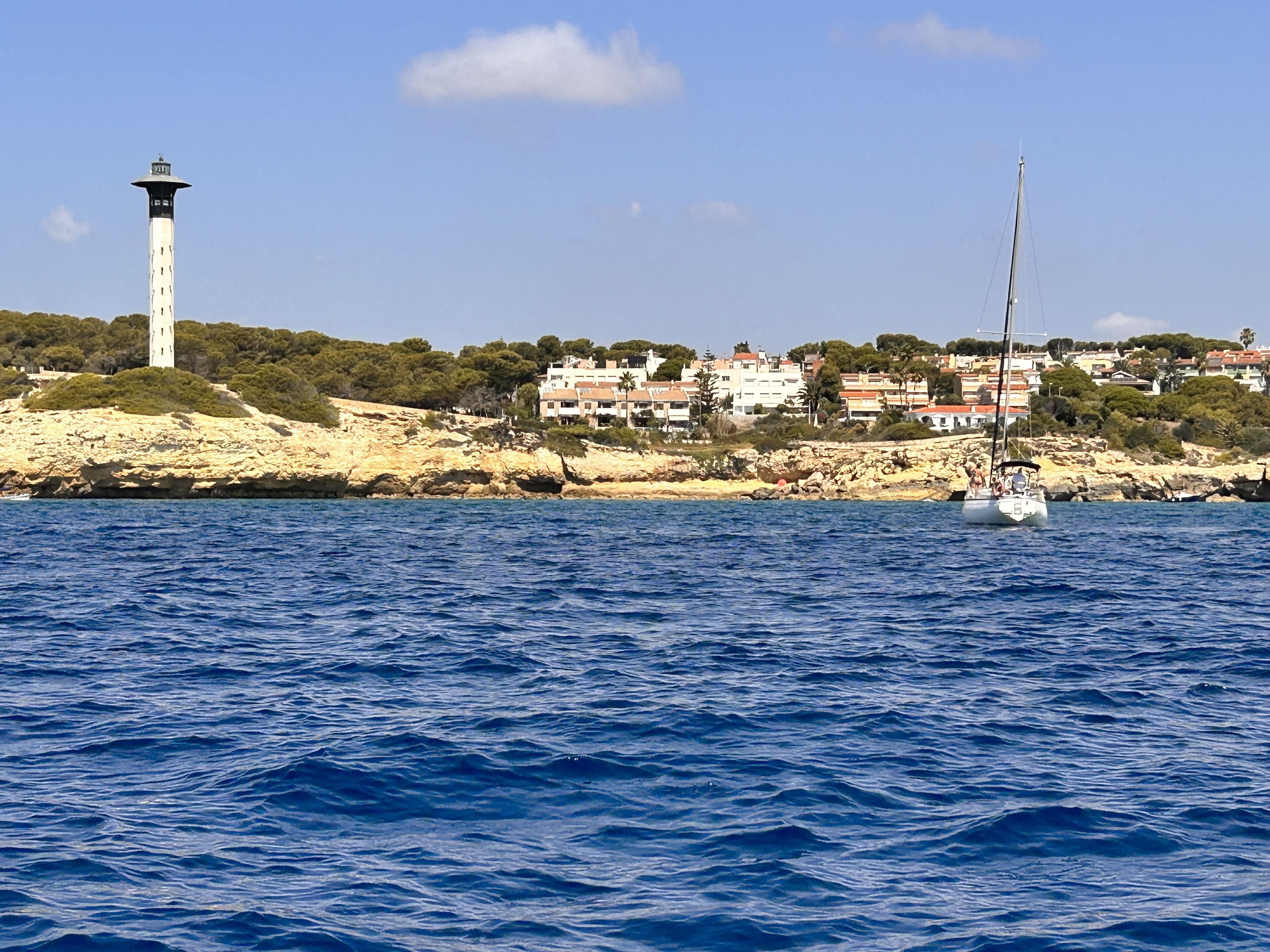 Un paysage côtier avec un phare, un voilier et des bâtiments le long de la côte.