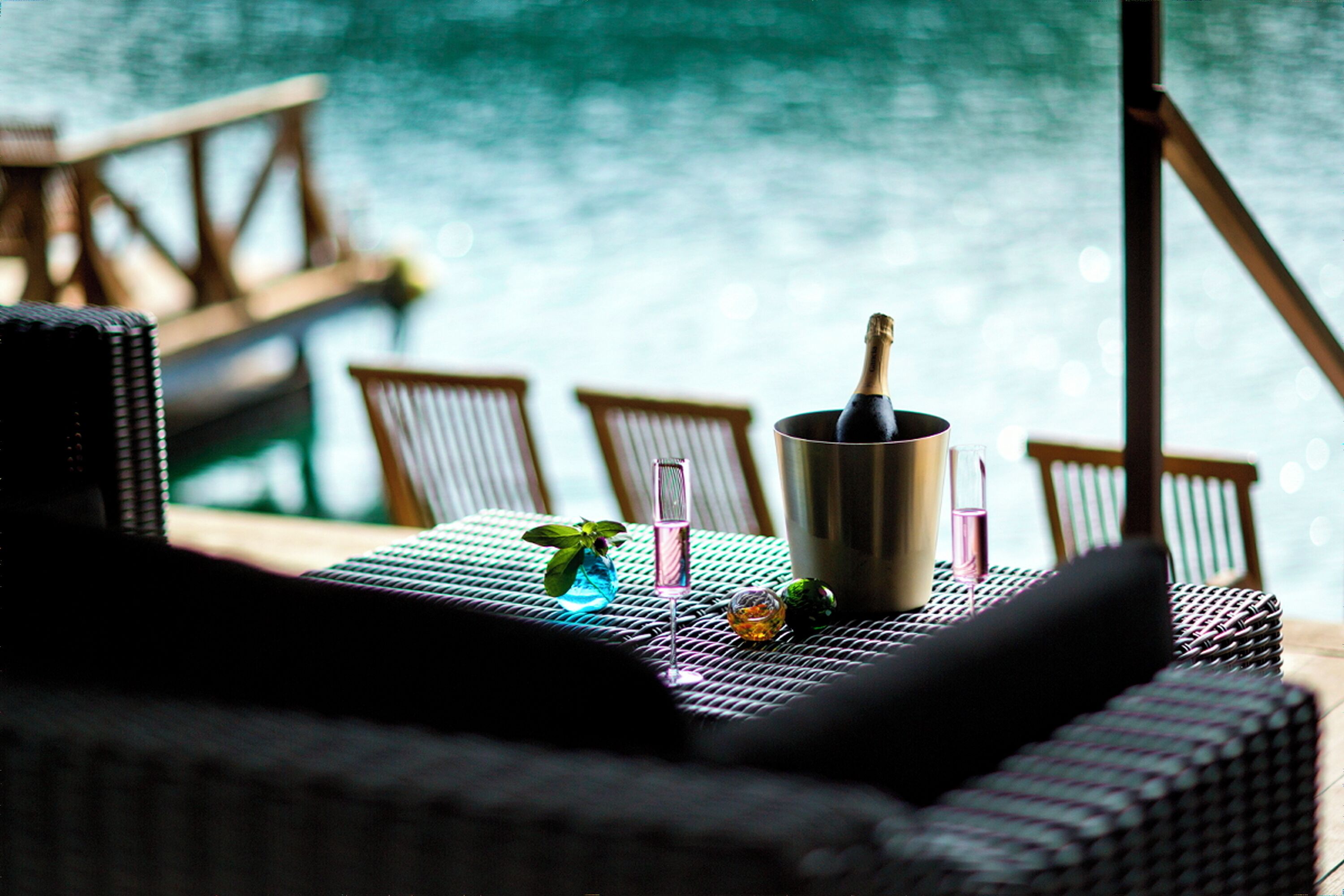Une chaise en osier, une bouteille de champagne et des verres sur une table placée au bord d’une piscine.