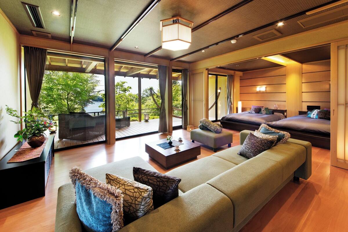 A modern hotel room with a large bed, a sofa, a coffee table, and a view of greenery outside.