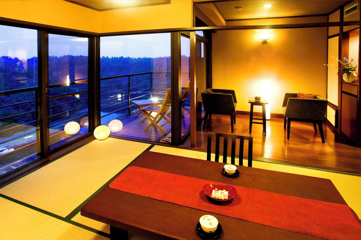 A traditional Japanese room with tatami flooring, a low table with a red runner, and a view of a balcony with a railing.