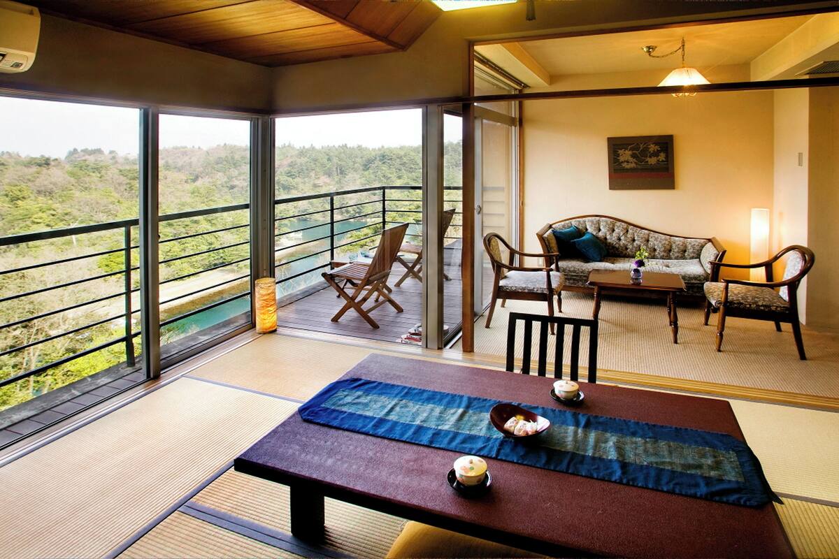 A room with a view of a forested area, a sofa, a dining table with a placemat, and a chair.