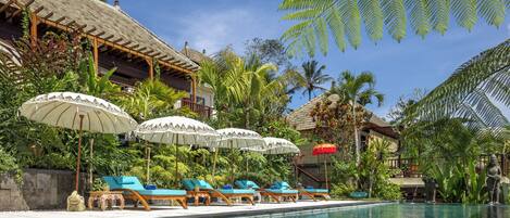 A pool area with lounge chairs and umbrellas, surrounded by tropical plants and a thatched-roof building.