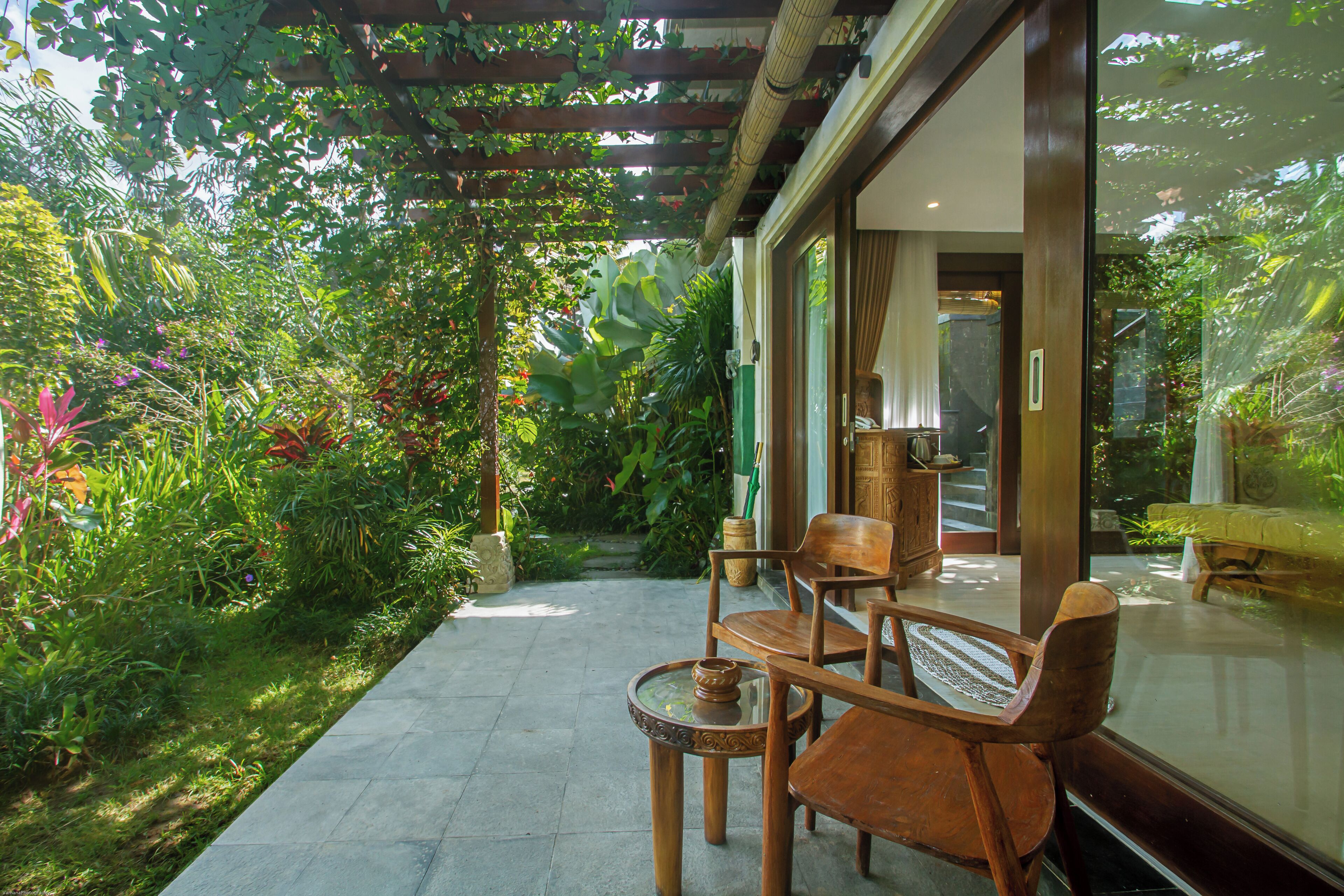 Un patio con mesa y sillas de madera, rodeado de abundante vegetación y una puerta de cristal que da a una habitación.
