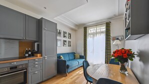 A modern kitchen with grey cabinets, a wooden countertop, and a built-in oven. There is a blue sofa with decorative pillows, a dining table with chairs, and a vase with red flowers. - La Maison Kléber – Luxury 1BR near Eiffel Tower & Champs-Élysées (Paris)