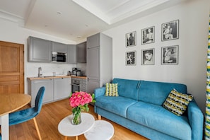 A modern living room with a blue sofa, a wooden dining table, and a kitchen in the background. - La Maison Kléber – Luxury 1BR near Eiffel Tower & Champs-Élysées (Paris)