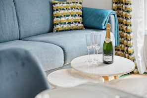 Sala moderna com um sofá azul, almofadas estampadas e uma pequena mesa branca com uma garrafa de champanhe e copos.