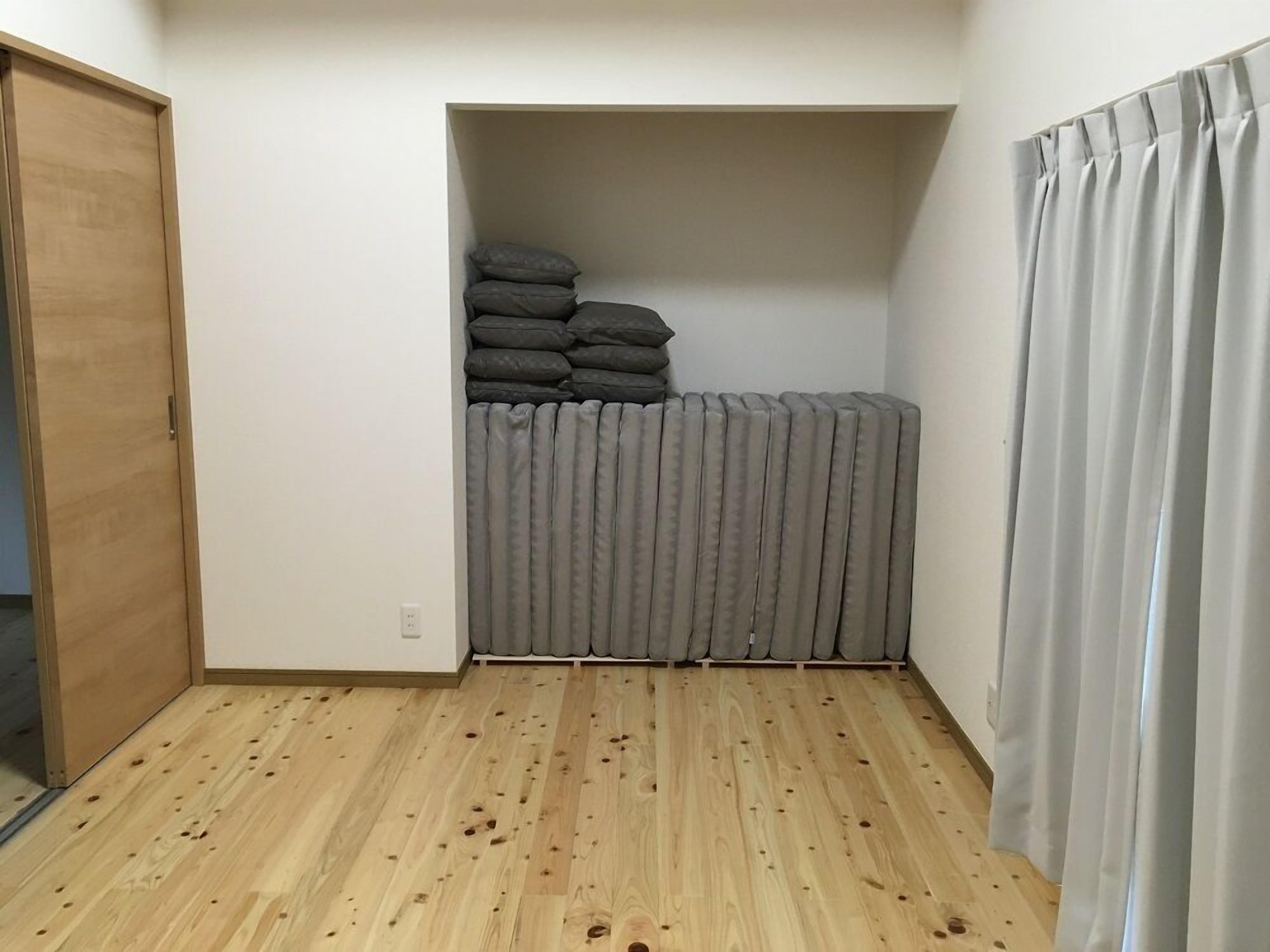 A room with a wooden floor, a radiator, a grey curtain, and a stack of pillows against the wall.