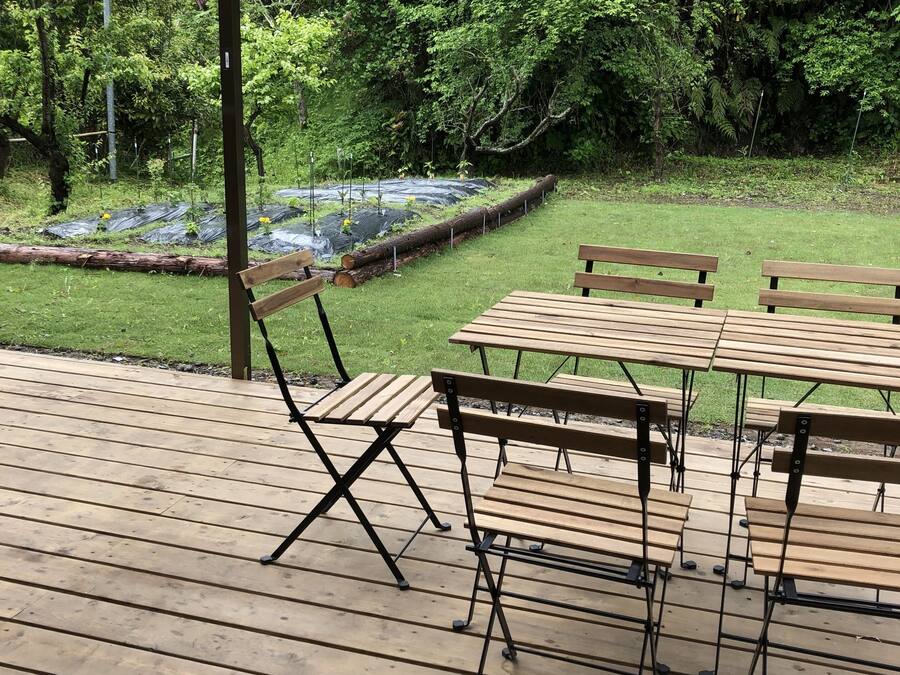 Una terraza de madera al aire libre con mesas y bancos, rodeada por un jardín con bancales elevados y una valla de madera.