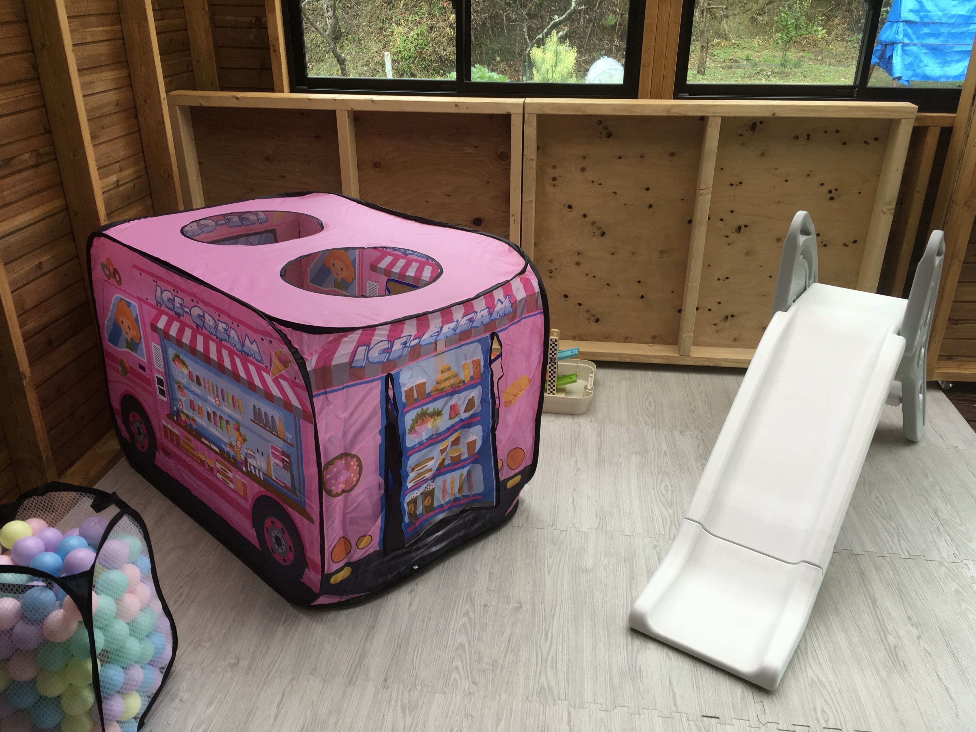 Una casita tipo camión de helados rosa con un tobogán, una piscina de pelotas y una ventana con vista a los árboles.