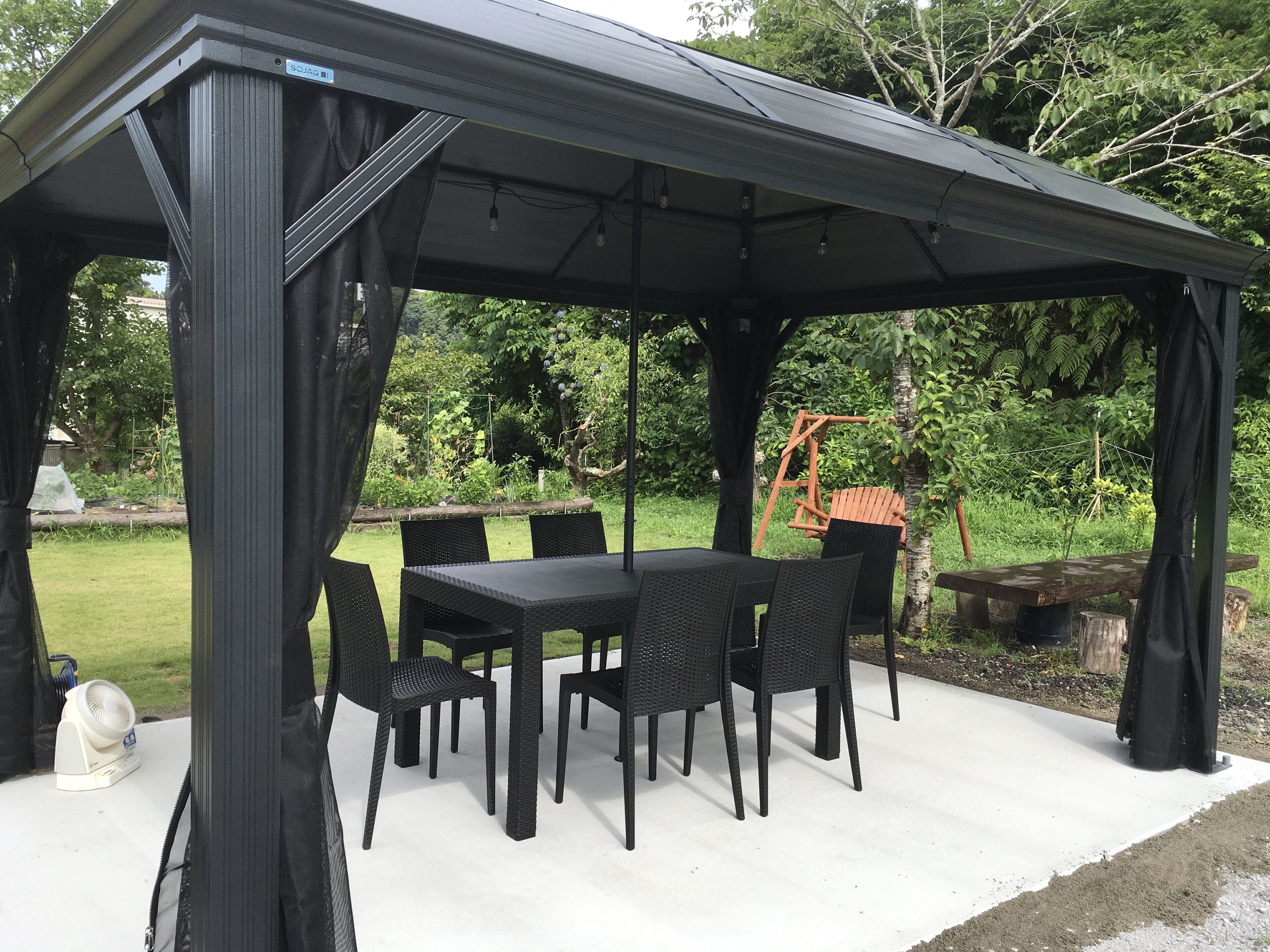 A covered outdoor patio with a table and chairs, surrounded by greenery.