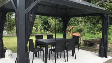 A covered outdoor patio with a table and chairs, surrounded by greenery.