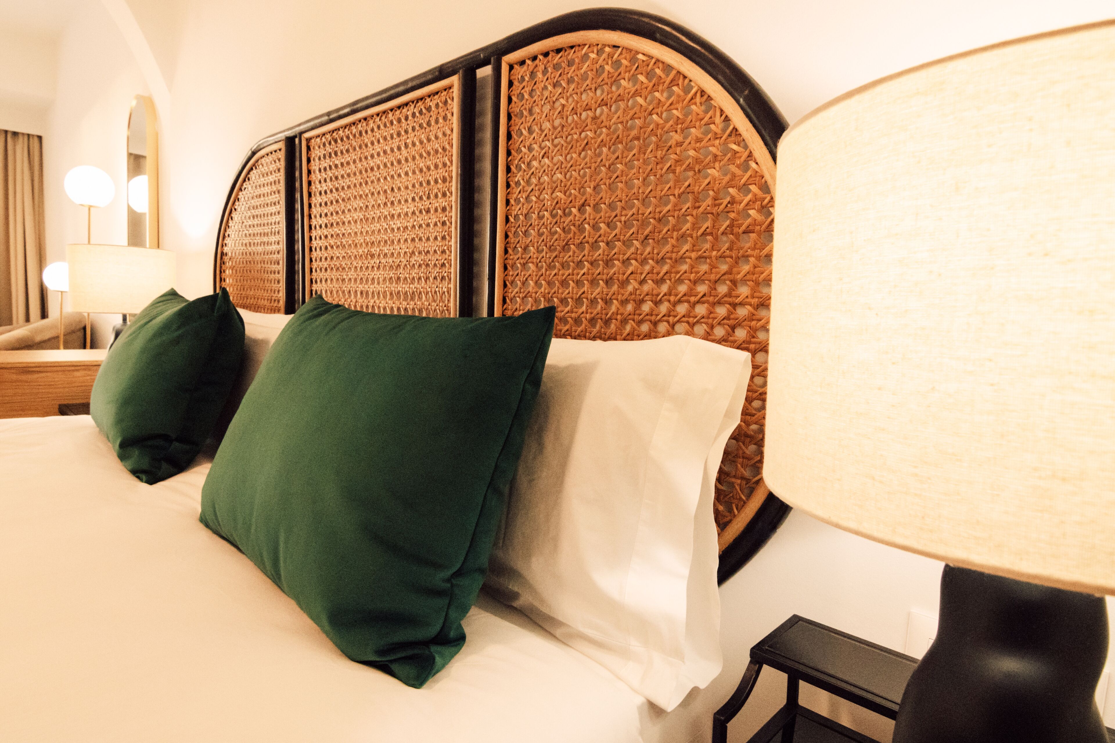 A neatly made bed with green and white pillows, a woven headboard, and a bedside lamp.