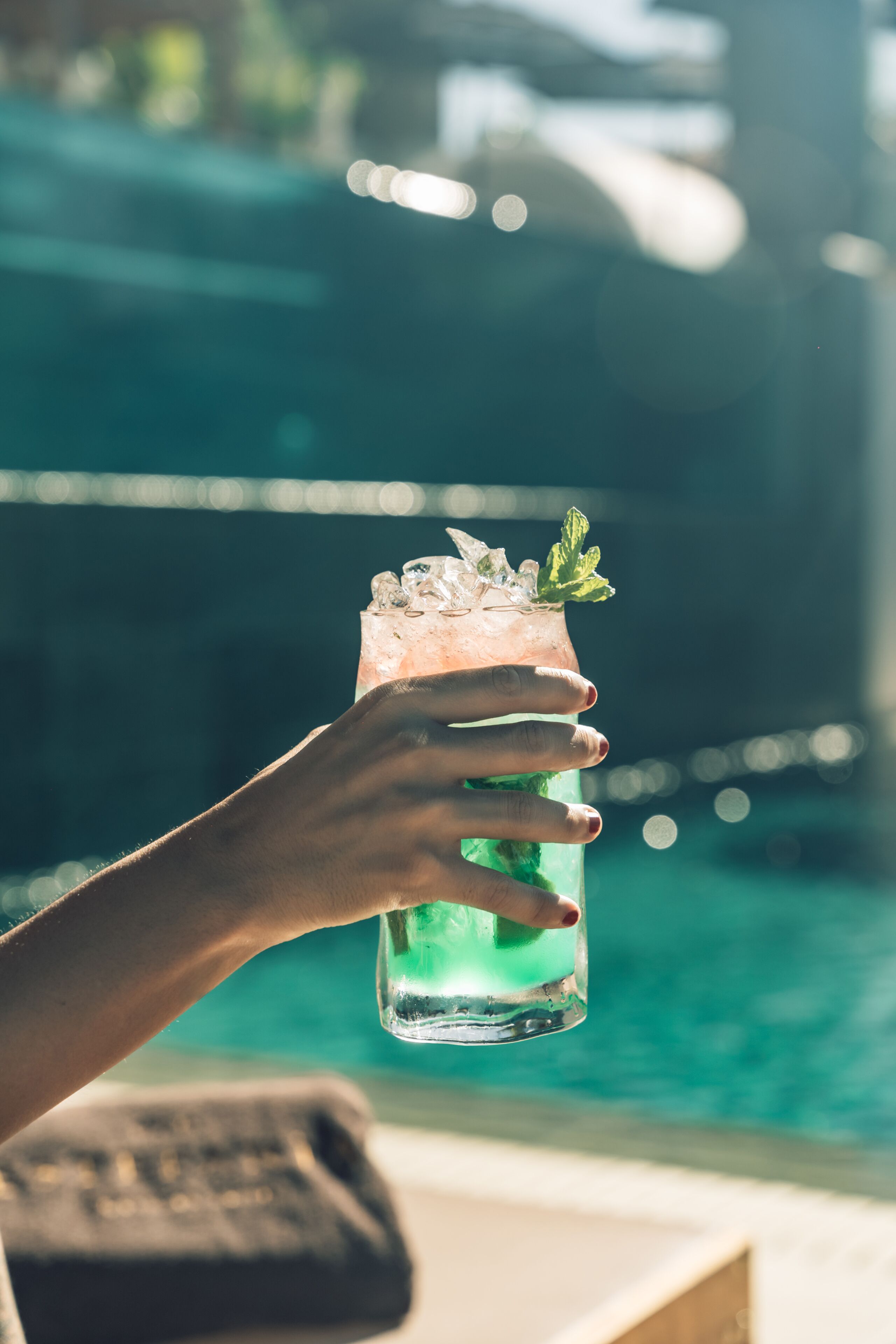 A hand holding a tall glass with a green drink, ice, and mint garnish, with a pool in the background.
