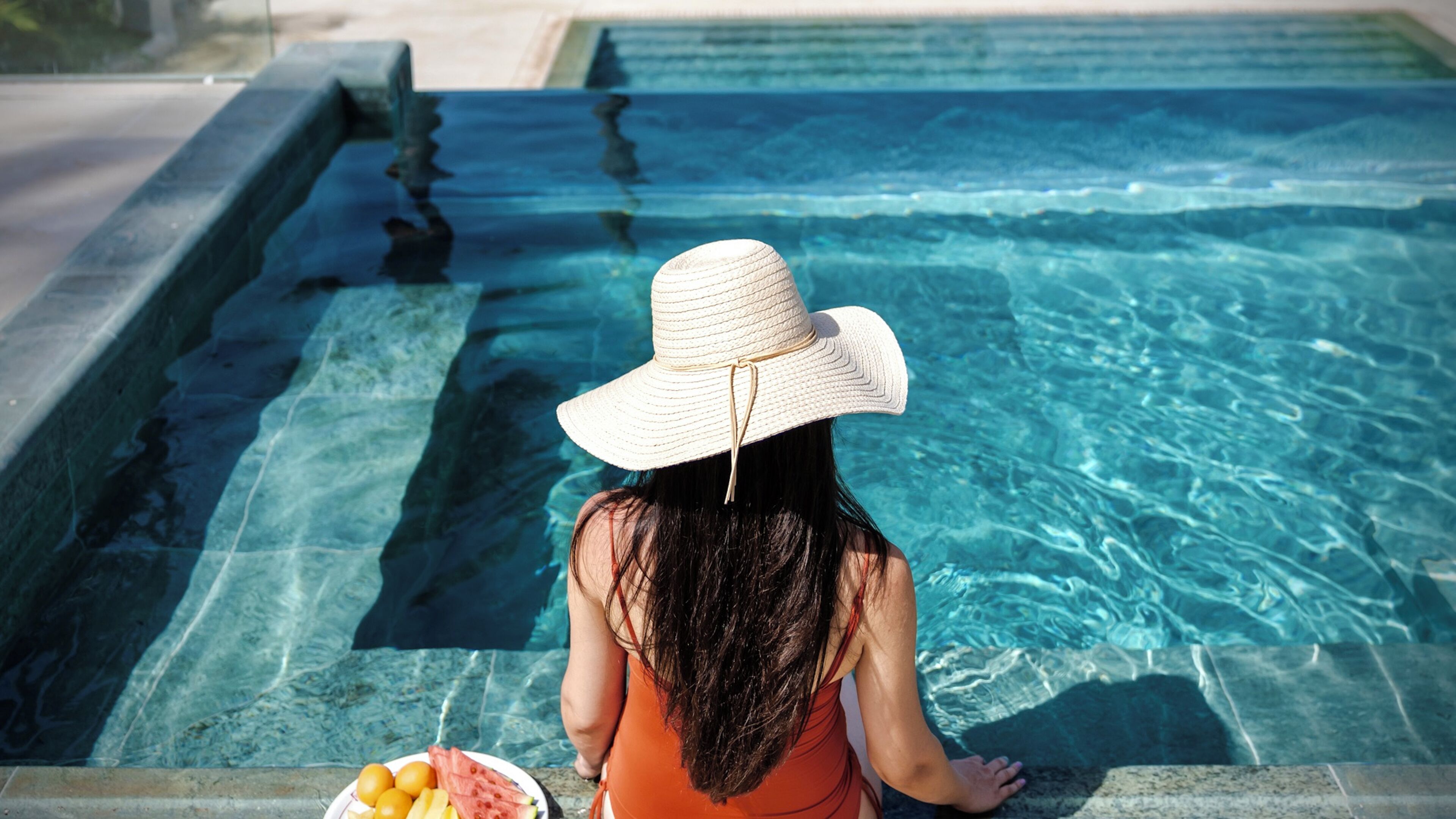 A person sitting by a pool with clear water, wearing a straw hat and an orange top.