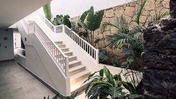 Uma escadaria com corrimãos brancos leva a um patamar, cercado por plantas tropicais e um muro de pedra.
