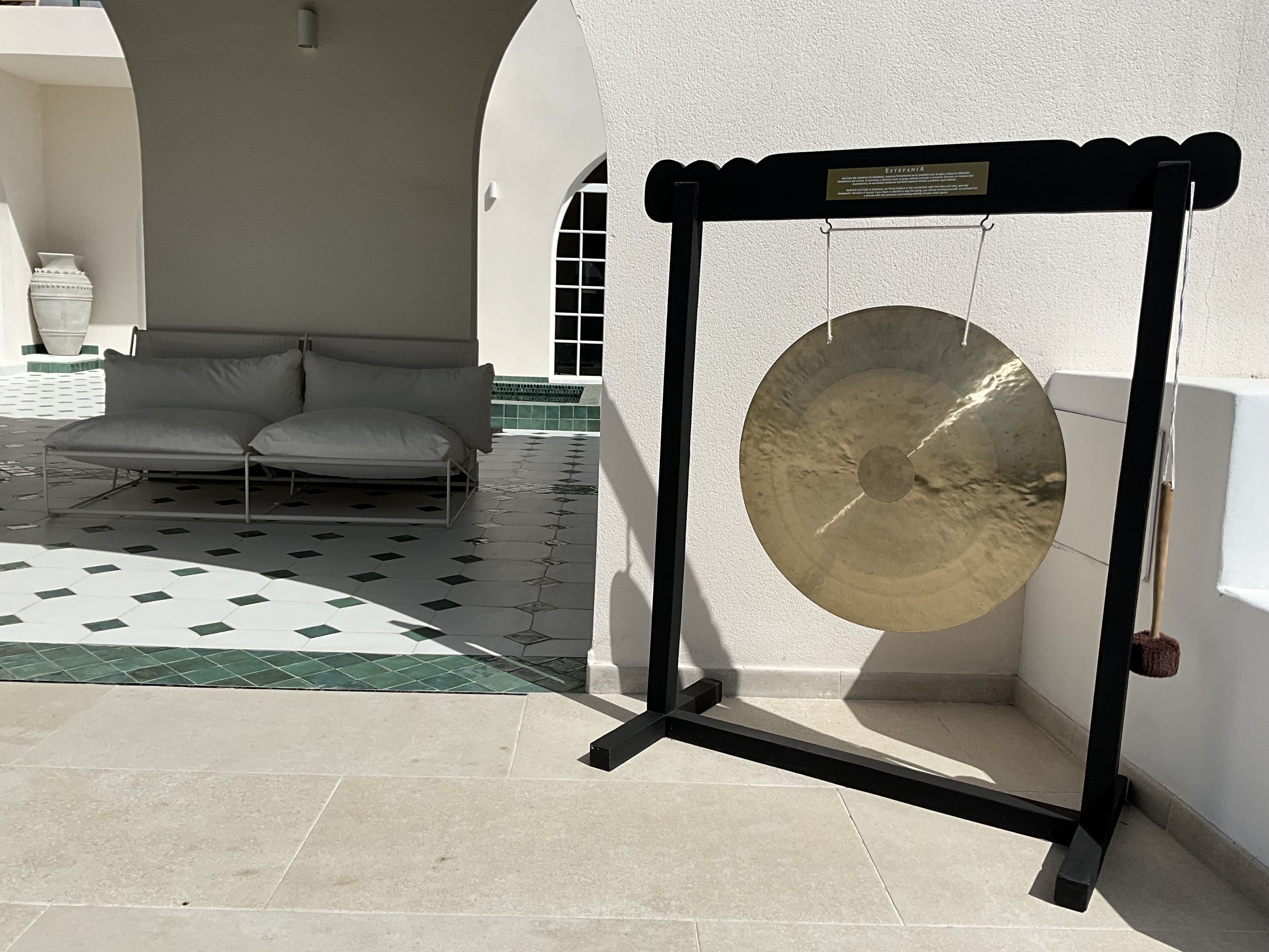 A room with a large gong mounted on a stand, a sofa with cushions, and a tiled floor with a geometric pattern.