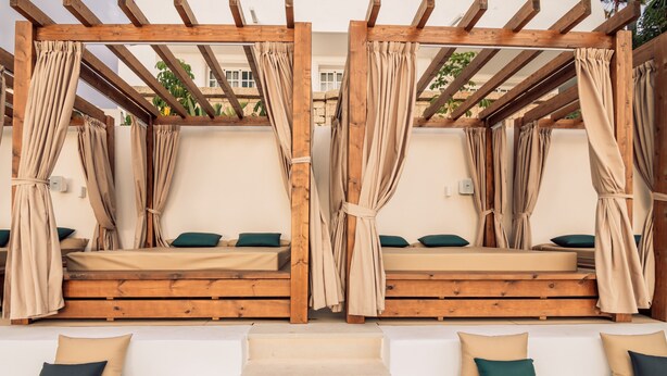 Twee houten bedden met beige beddengoed en groene kussens, onder een houten pergola met beige gordijnen.