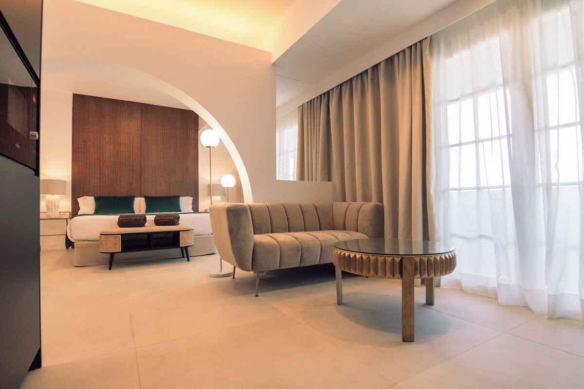 Een moderne hotelkamer met een bank, een glazen salontafel, een bed met hoofdbord en een groot raam met gordijnen.