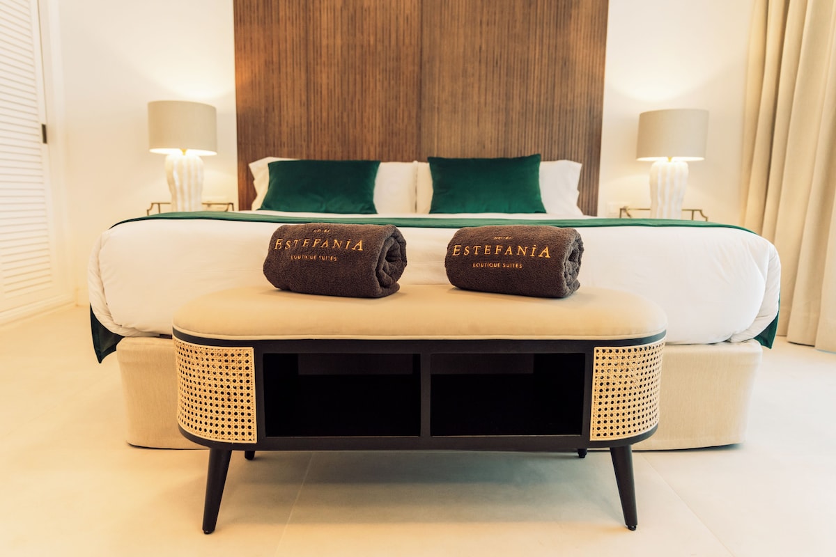 Une chambre d’hôtel comprenant un lit, deux serviettes aux couleurs de « Estefania Boutique Suites », une table de chevet et deux lampes.