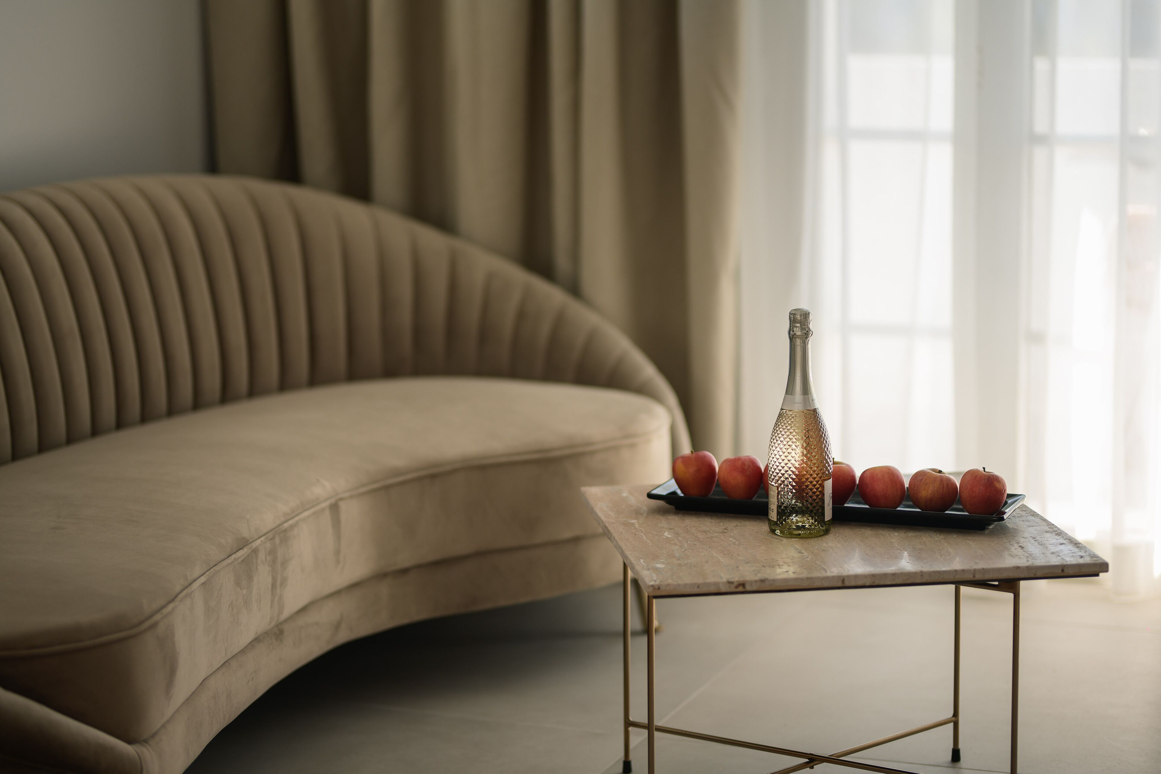 Un canapé beige, une petite table avec une bouteille et des pommes, et une fenêtre avec des rideaux.