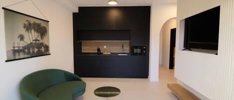 Een moderne woonkamer met een groene bank, een klein rond tafeltje en een keukenhoek met een zwart aanrecht en een houten achterwand.