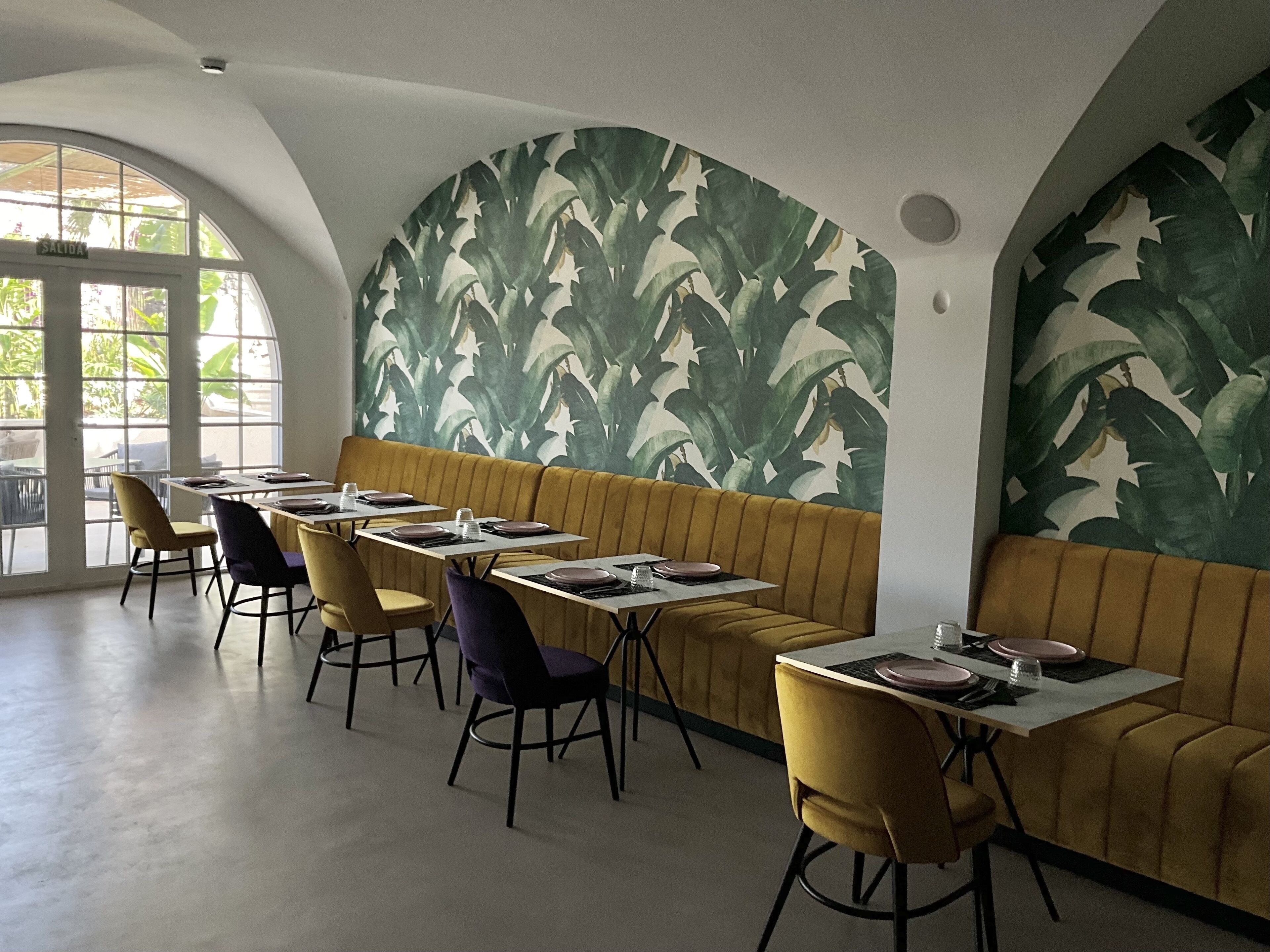 Un restaurante con una larga zona de asientos tipo banco, mesas con sillas y un amplio ventanal que proporciona luz natural.