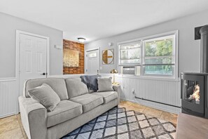 Ein modernes Wohnzimmer mit einem grauen Sofa, einem gemusterten Teppich, einem Kamin und großen Fenstern mit Jalousien.