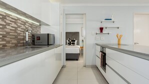 Una cocina moderna con horno empotrado, microondas y cafetera. Se aprecia una cama con cabecera gris y almohada blanca y negra. La cocina cuenta con gabinetes blancos y salpicadero de azulejos.