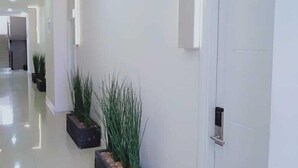 A modern, well-lit hallway with white walls and a tiled floor. There are two potted plants and a door on the right side. - DAIMA Syariah Mansion Tebet (Jakarta)