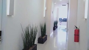 A clean, modern hallway with white walls and a shiny tiled floor. There are potted plants on the left and a fire extinguisher on the right. - DAIMA Syariah Mansion Tebet (Jakarta)