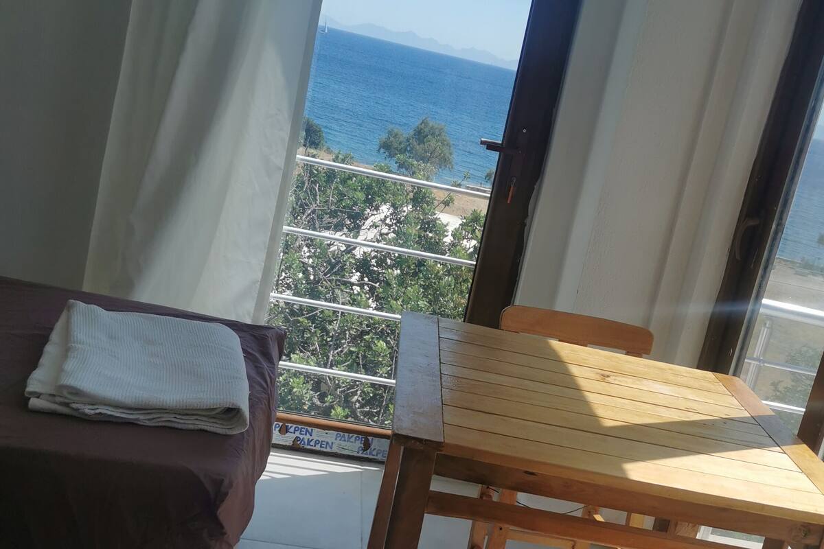 Varanda com mesa e cadeiras de madeira, cama com cobertor branco e vista para o mar e montanhas.