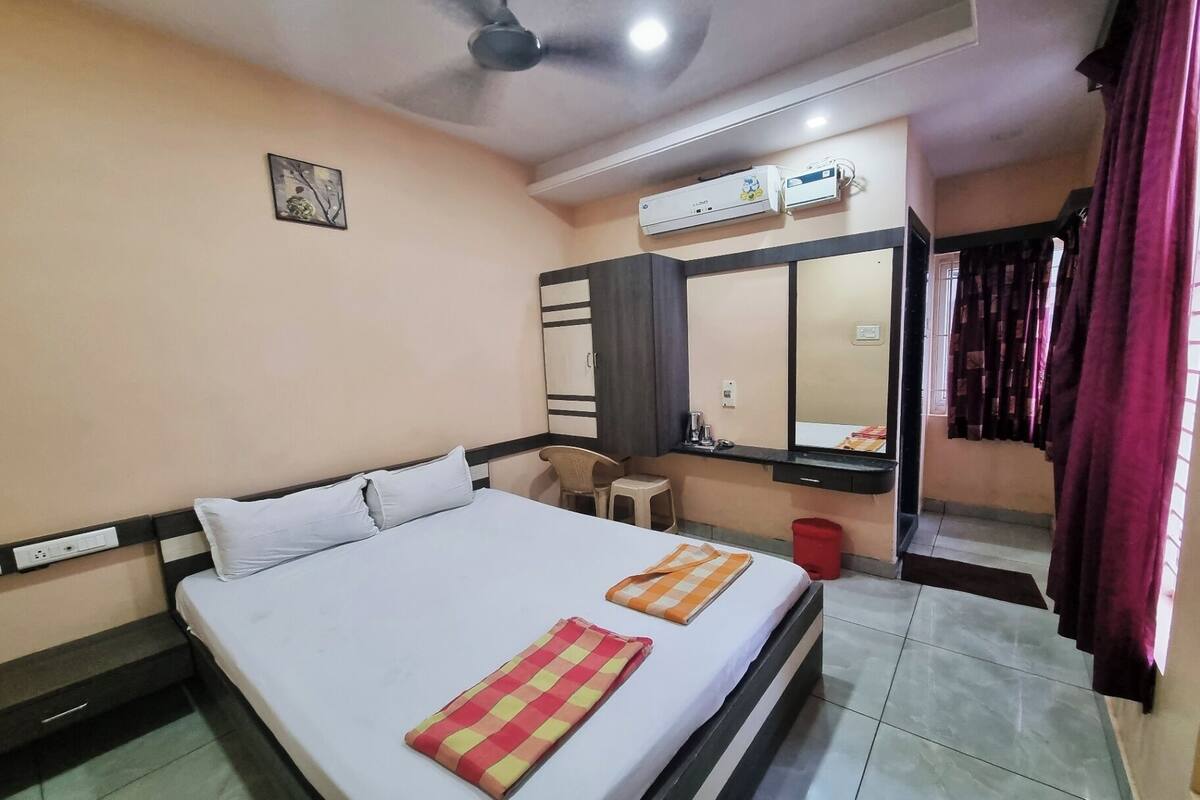 A hotel room with a bed, a desk, a chair, a mirror, and an air conditioning unit.