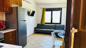 A compact kitchen with a refrigerator, stove, and a flat-screen TV. A gray sofa and a dining table with a chair are in the living area. - Cute Apple - At The Heart Of The Old Town (Chania)