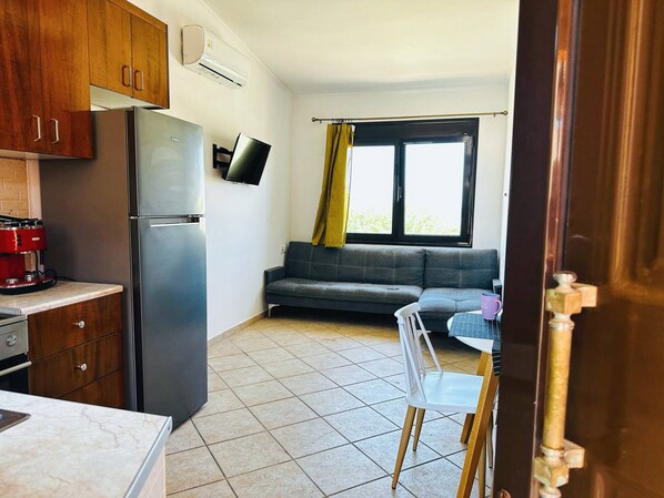 Una cocina compacta con refrigerador, estufa y un televisor de pantalla plana. Un sofá gris y una mesa de comedor con una silla están en la sala.