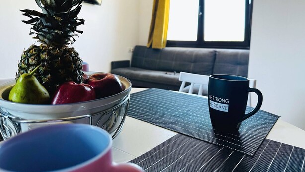 A living room with a glass bowl containing fruits, a black mug with text, and a window with curtains. - Cute Apple - At The Heart Of The Old Town (Chania)