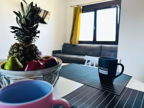 Un salón con un cuenco de vidrio con frutas, una taza negra con texto y una ventana con cortinas.