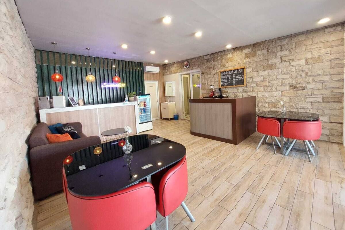 A modern hotel lobby with a reception desk, a seating area with a glass-top table and red chairs, and a stone wall.
