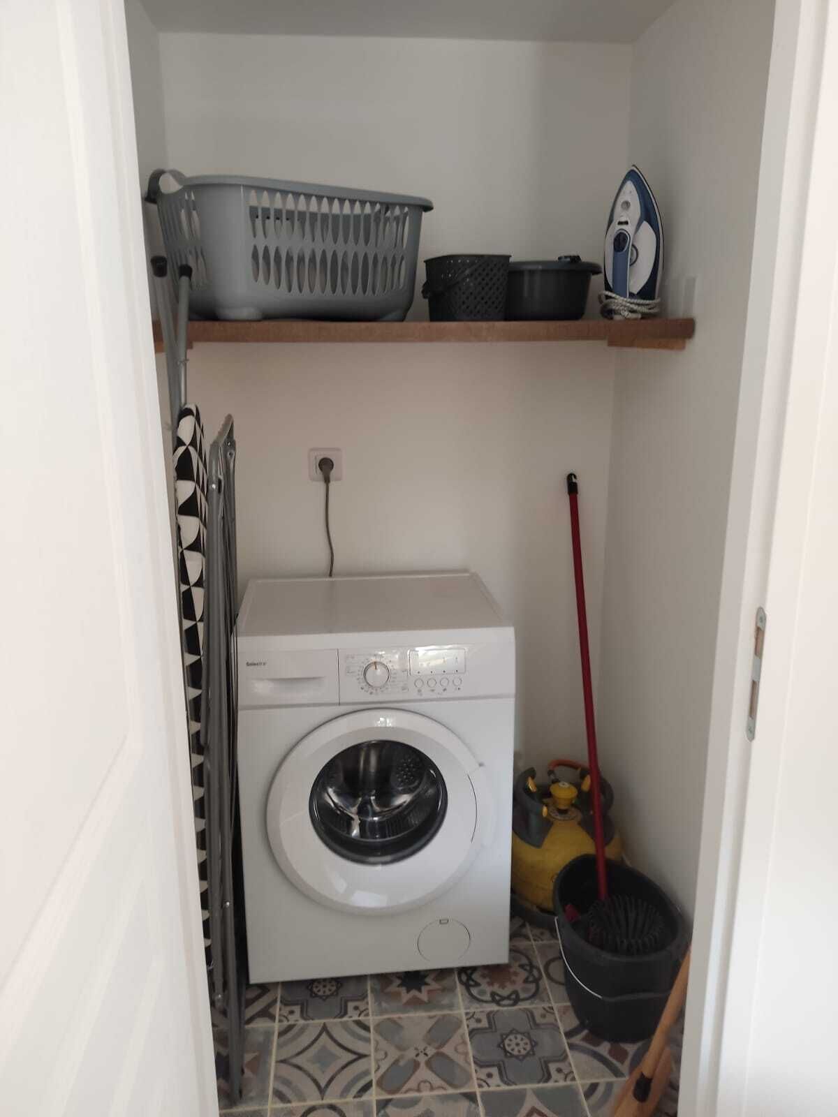 Une petite buanderie comprenant un lave-linge, un étendoir et une étagère où sont rangés les produits de lessive.