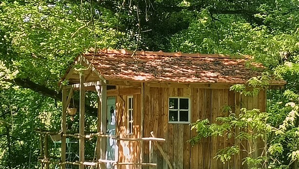 Una casa del árbol de madera con techo de tejas rojas, construida sobre pilares, rodeada de follaje denso.