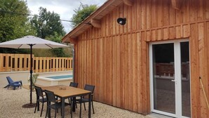 Un edificio de madera con piscina, un porche cubierto con mesa y sillas, y una valla de madera al fondo.