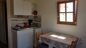 A compact kitchen with a coffee maker, a sink, and a small refrigerator. There is a window with a view of buildings outside. - Joli Petit Appartement Neuf, au Calme et à Proximité de la mer et des Commerces (La Guérinière)