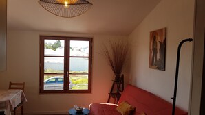 A room with a large window, a red sofa, a small table with a bowl, a chair, a lamp, and a painting on the wall. - Joli Petit Appartement Neuf, au Calme et à Proximité de la mer et des Commerces (La Guérinière)