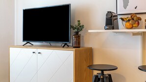 Ein modernes Wohnzimmer mit Fernseher auf einer Holzkommode, einem kleinen Tisch mit Pflanze und einer Obstschale auf einem Regal.