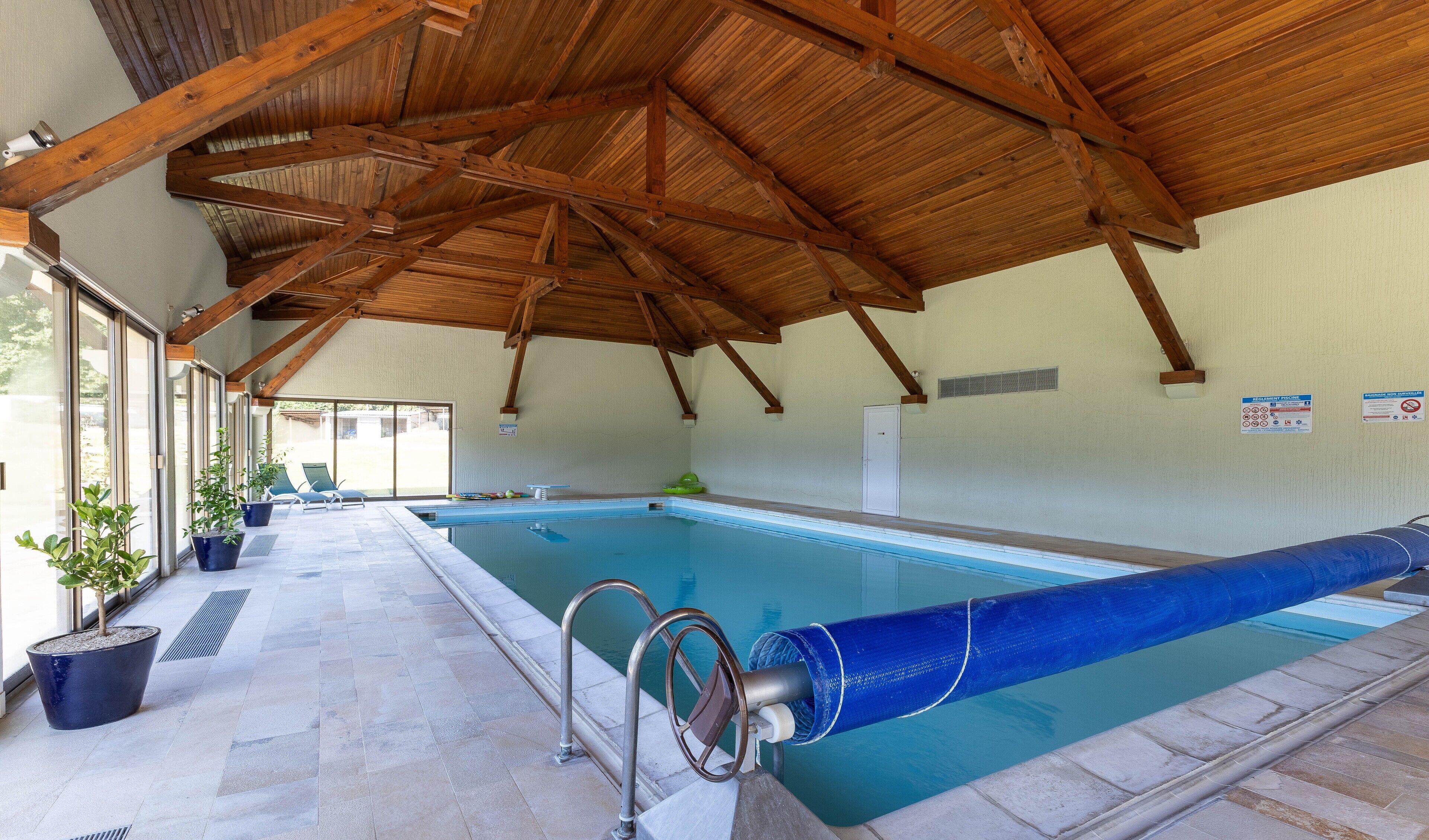 An indoor swimming pool with a wooden ceiling, clear glass walls, and a tiled floor.