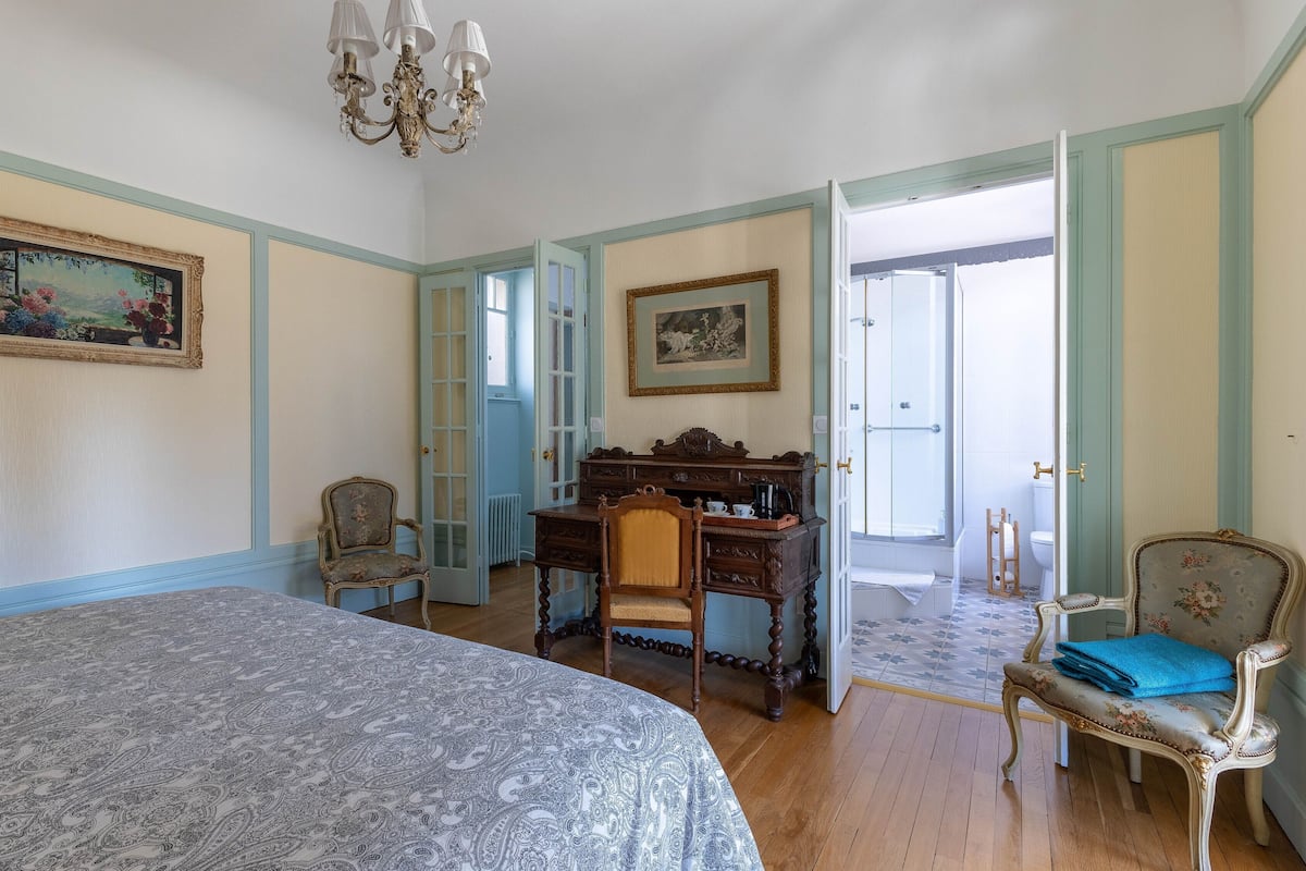 Un dormitorio con una cama grande, un escritorio con una silla, una silla con un cojín azul y una puerta que da a un baño.