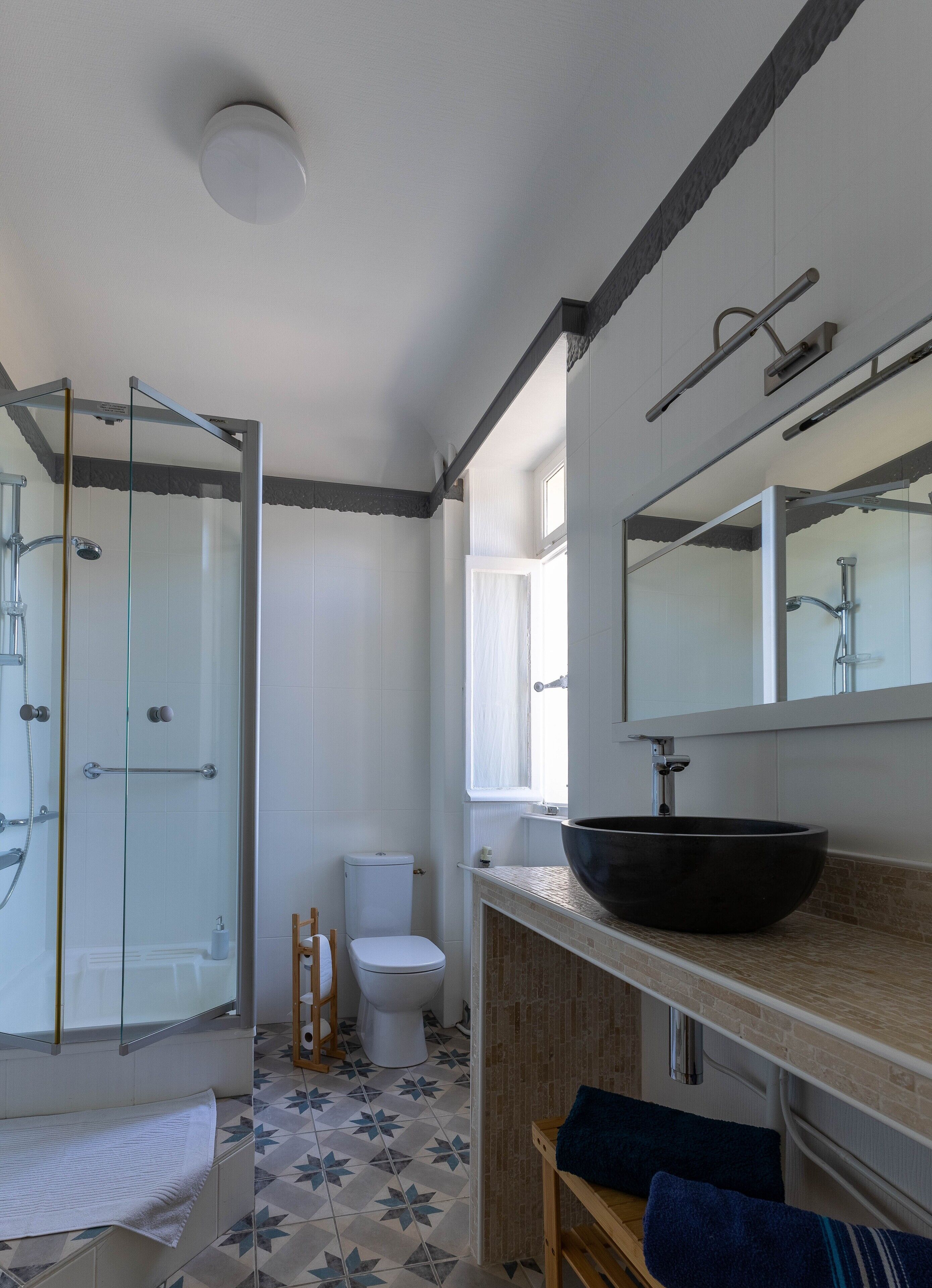 A bathroom with a glass shower enclosure, a white toilet, a large black vessel sink, and a wooden bench.