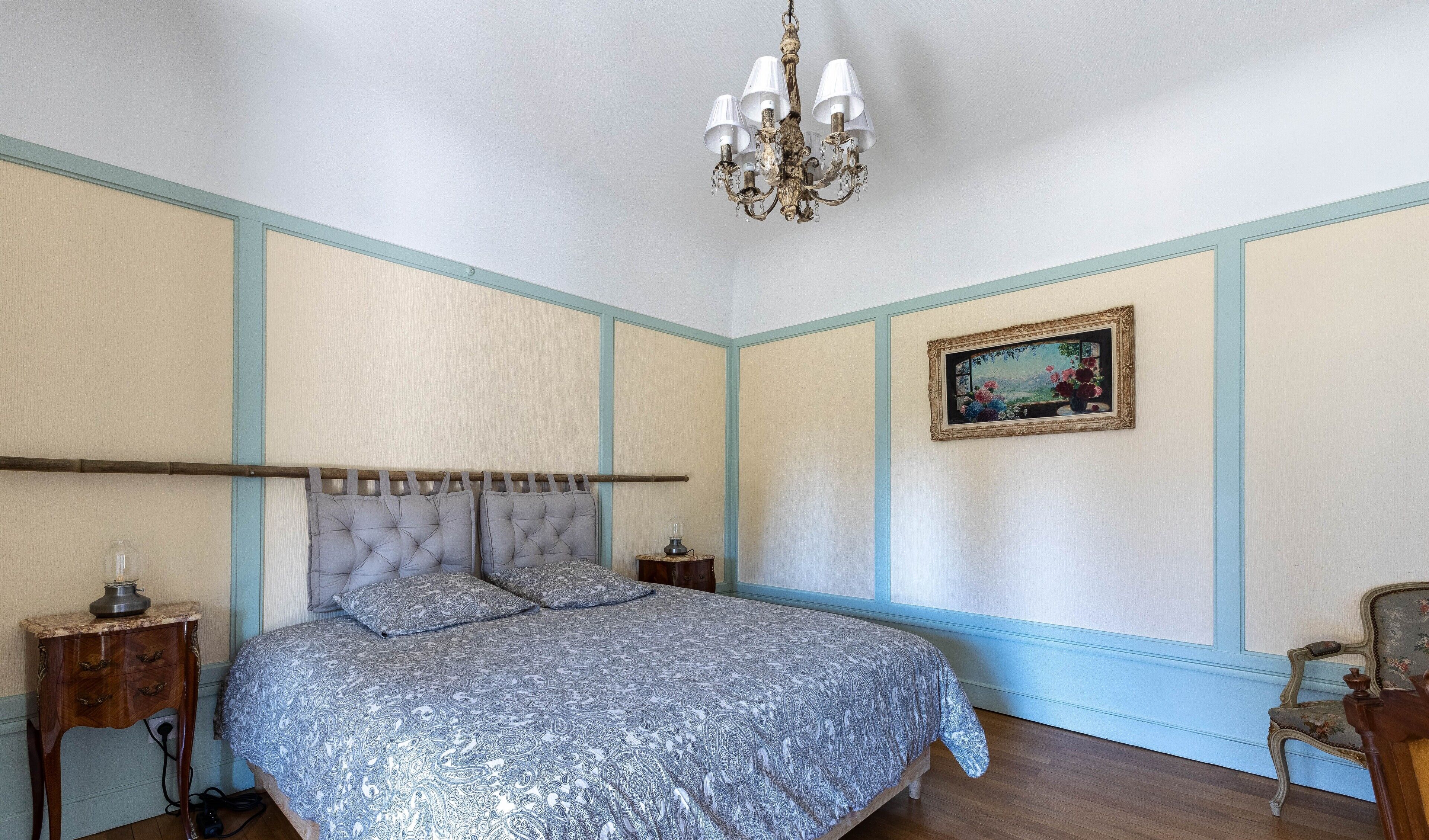 Une chambre à coucher comprenant un lit, un lustre, une table de chevet, une lampe, un tableau encadré et une chaise.